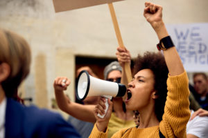 A young Black woman holding a megaphone raises her fist and shouts. Behind her, a sign reads We Need Justice.