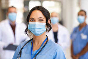 A healthcare worker in blue scrubs smiles at the camera behind their surgical mask.