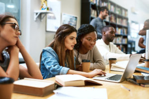 Two women sit together at a library, looking at a laptop, among other people reading and working.